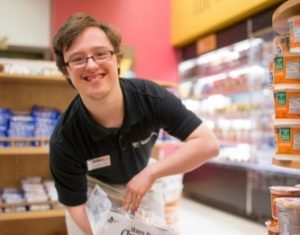 A young man smiles as he restocks items on the shelves in a grocery store.