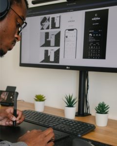 An African American man sits before a desktop computer with a large screen and uses a stylus pen and tablet to design a web page.