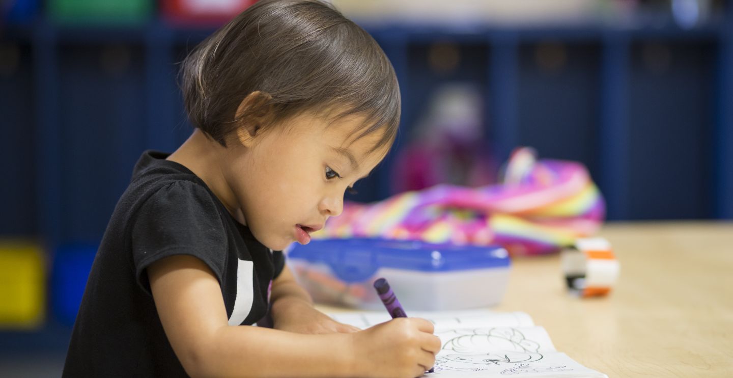 A very young girl in a classroom grasps a purple crayon and focuses completely on coloring the picture in her coloring book.