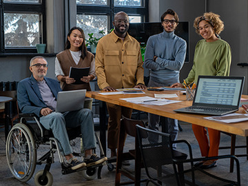 Six disability advocates, one of them seated in a wheelchair, smile as they pose next to a work table in a conference room
