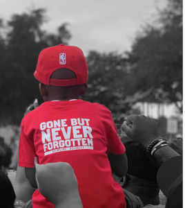 A young African American boy wearing a red tee shirt emblazoned with the words Gone But Never Forgotten rides on a man’s shoulders as they participate in a march in memory of Trayvon Martin.